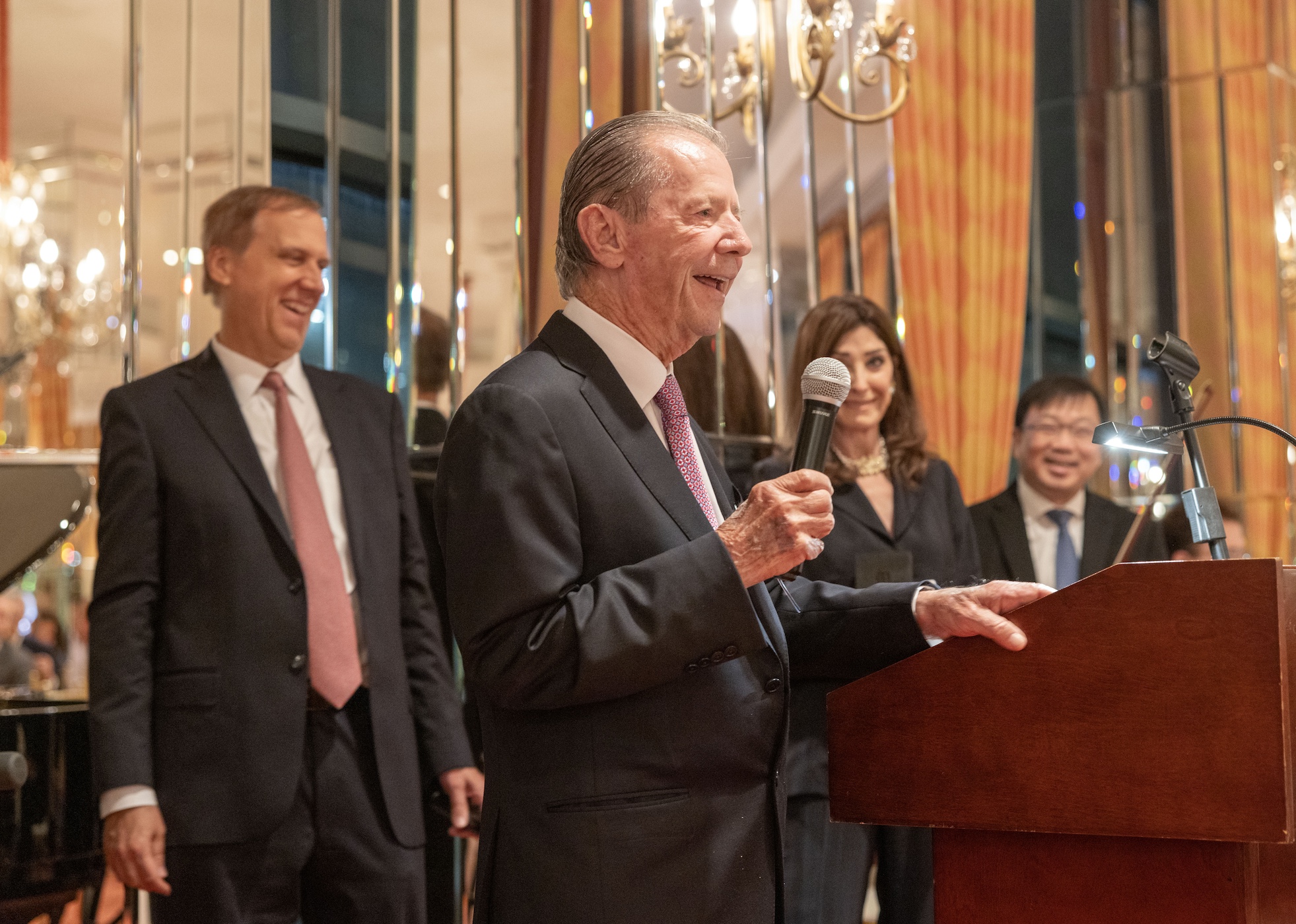 Elderly man holding microphone and smiling surrounded by smiling and clapping people