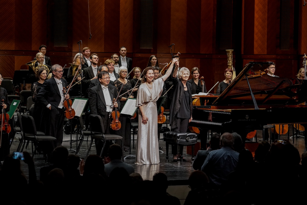 Elderly female conductor in all black with short hair holding raised hand of middle aged white woman with long brown hair and a silver dress in front of smiling orchestra musicians and piano
