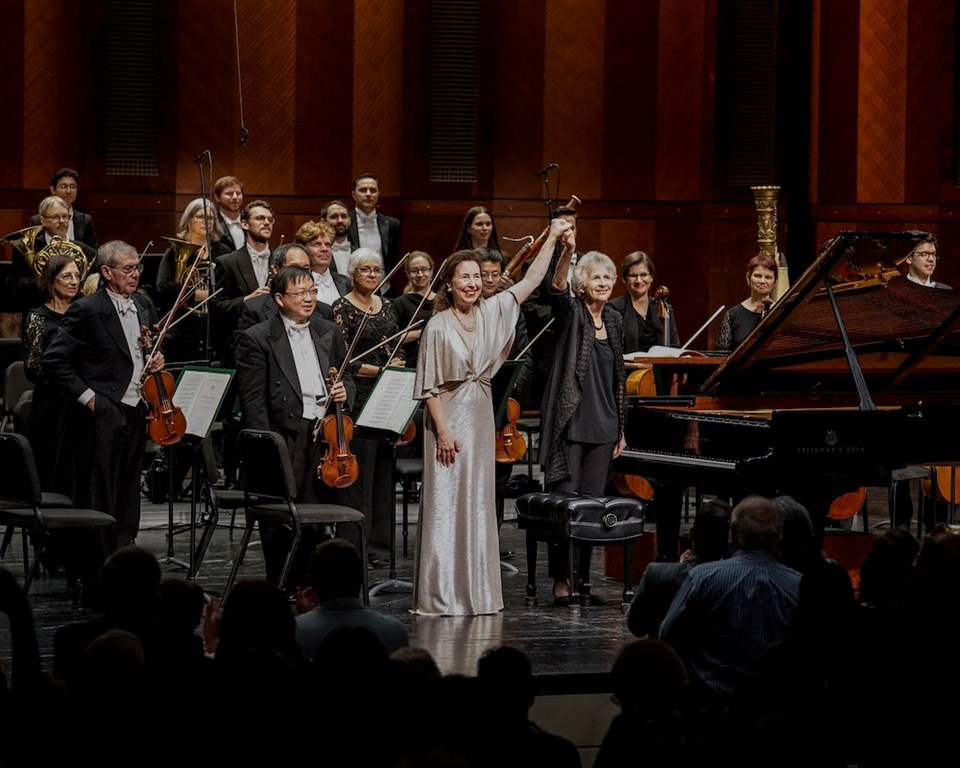 Elderly female conductor in all black with short hair holding raised hand of middle aged white woman with long brown hair and a silver dress in front of smiling orchestra musicians and piano