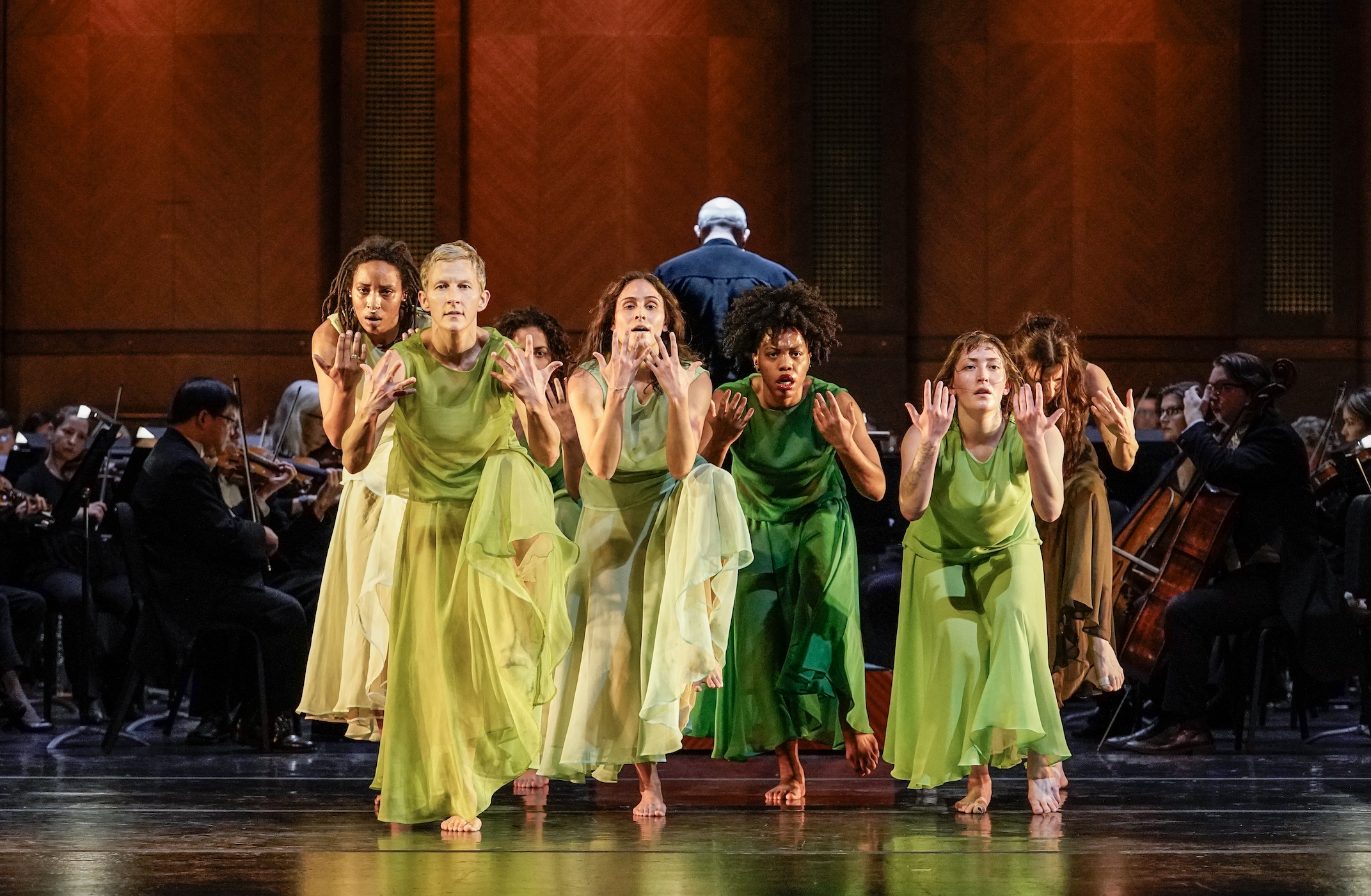 dancers in green dresses huddled together facing forward with one foot lifted and hands in front of their faces