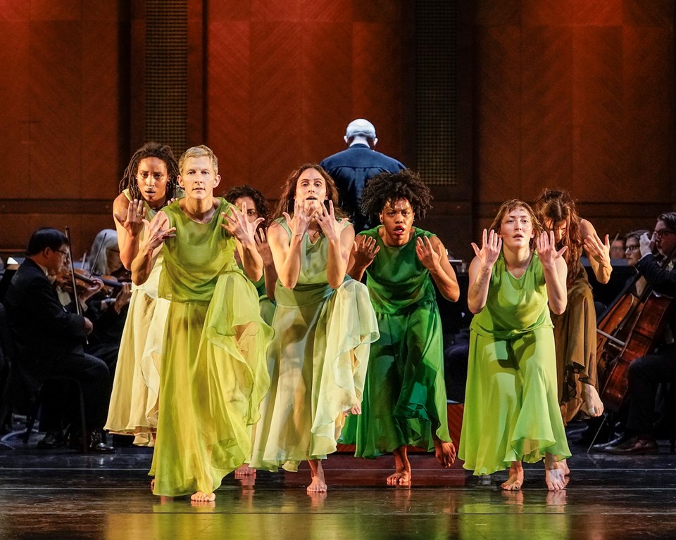 dancers in green dresses huddled together facing forward with one foot lifted and hands in front of their faces
