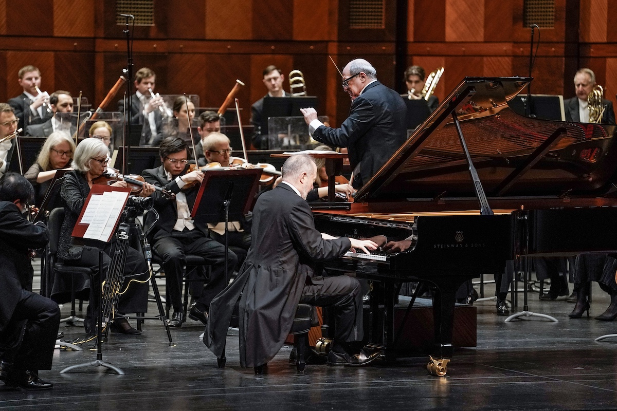 white man with grey hair wearing tails playing piano in front of full orchestra