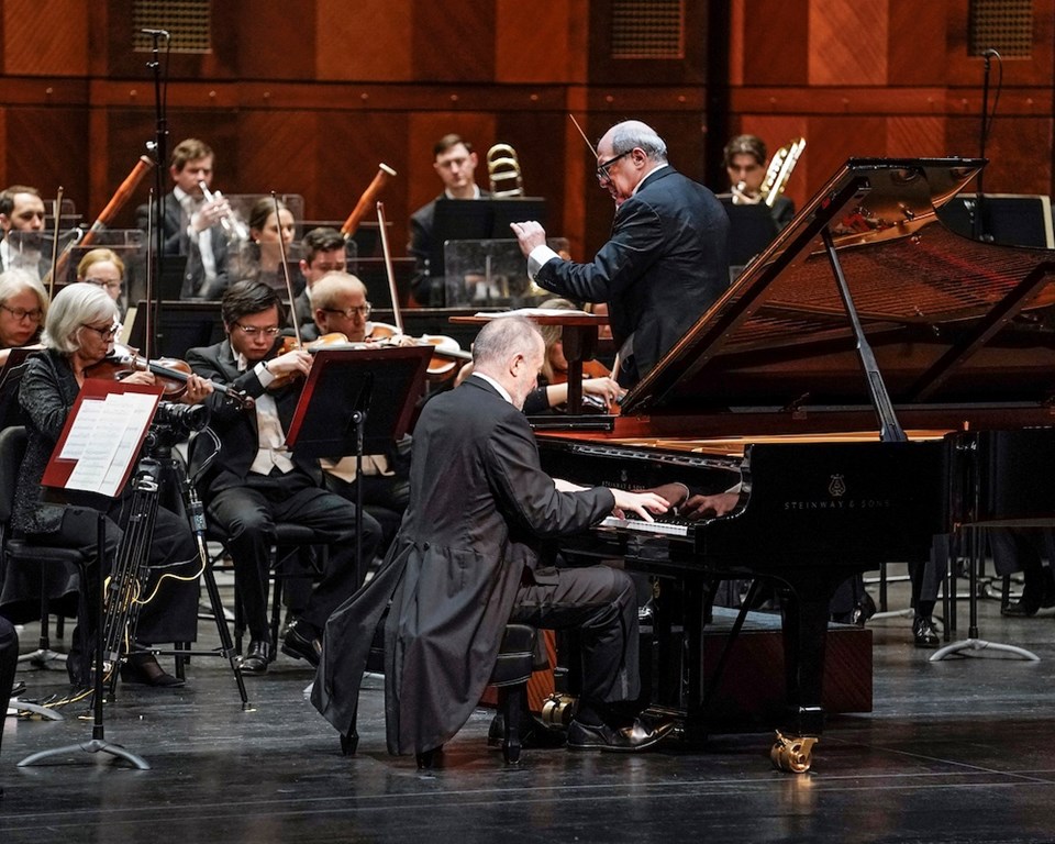 white man with grey hair wearing tails playing piano in front of full orchestra