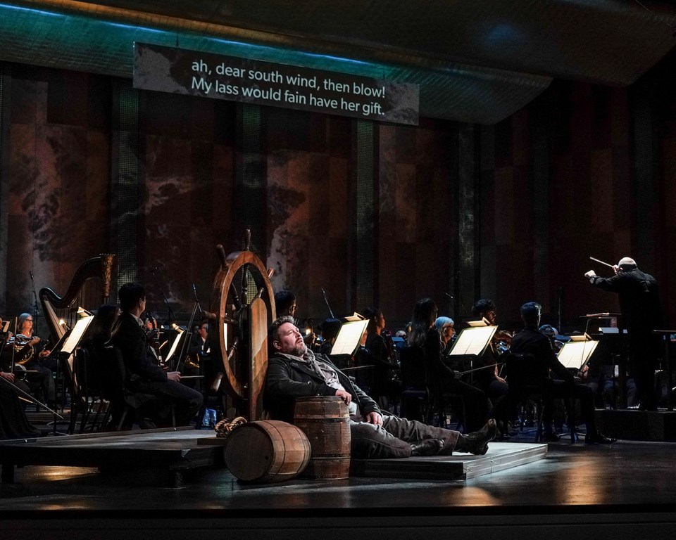 man opera singer leaning on ship wheel in front of orchestra