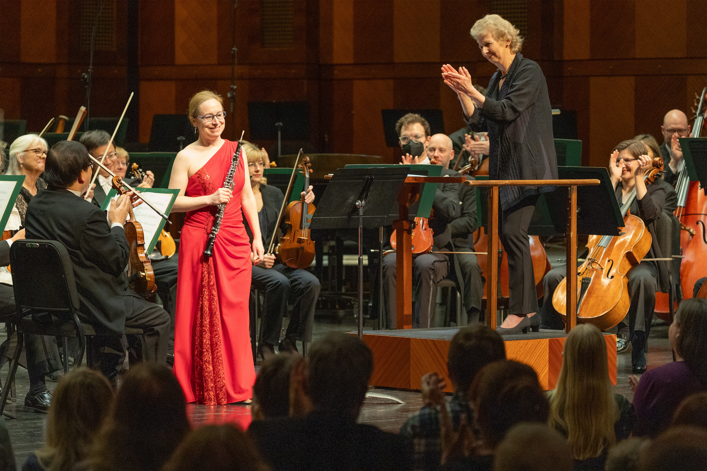 Female oboe player in red dress smiling on stage with conductor clapping for her