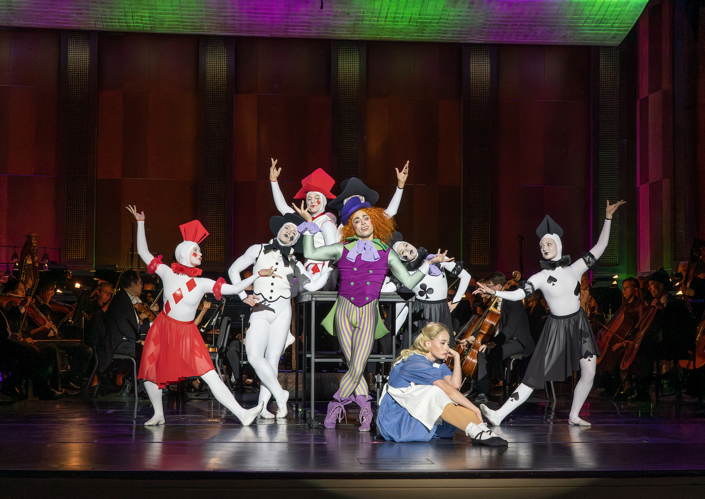 Alice in Wonderland dancers posing on stage on table in front of an orchestra