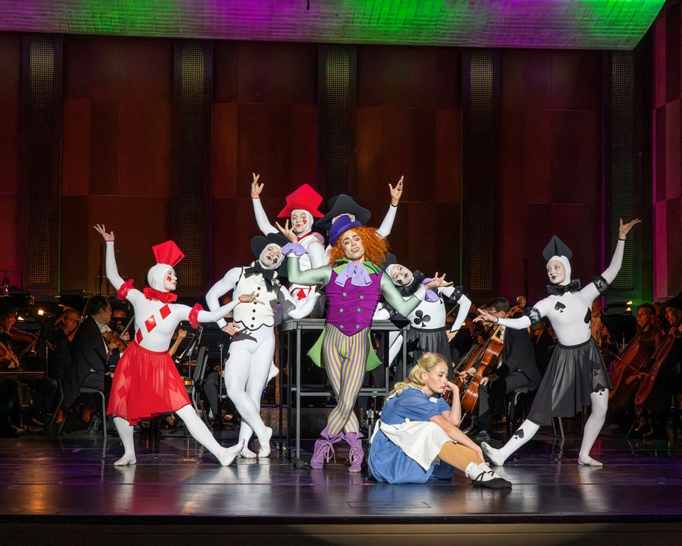 Alice in Wonderland dancers posing on stage on table in front of an orchestra